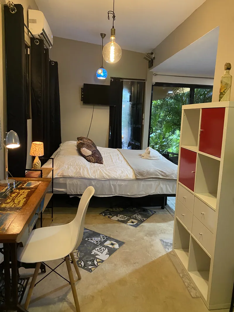 The studio bedroom at dusk with a made bed, a desk and lamp, a wall-mounted TV, and sliding glass doors opening to the garden.
